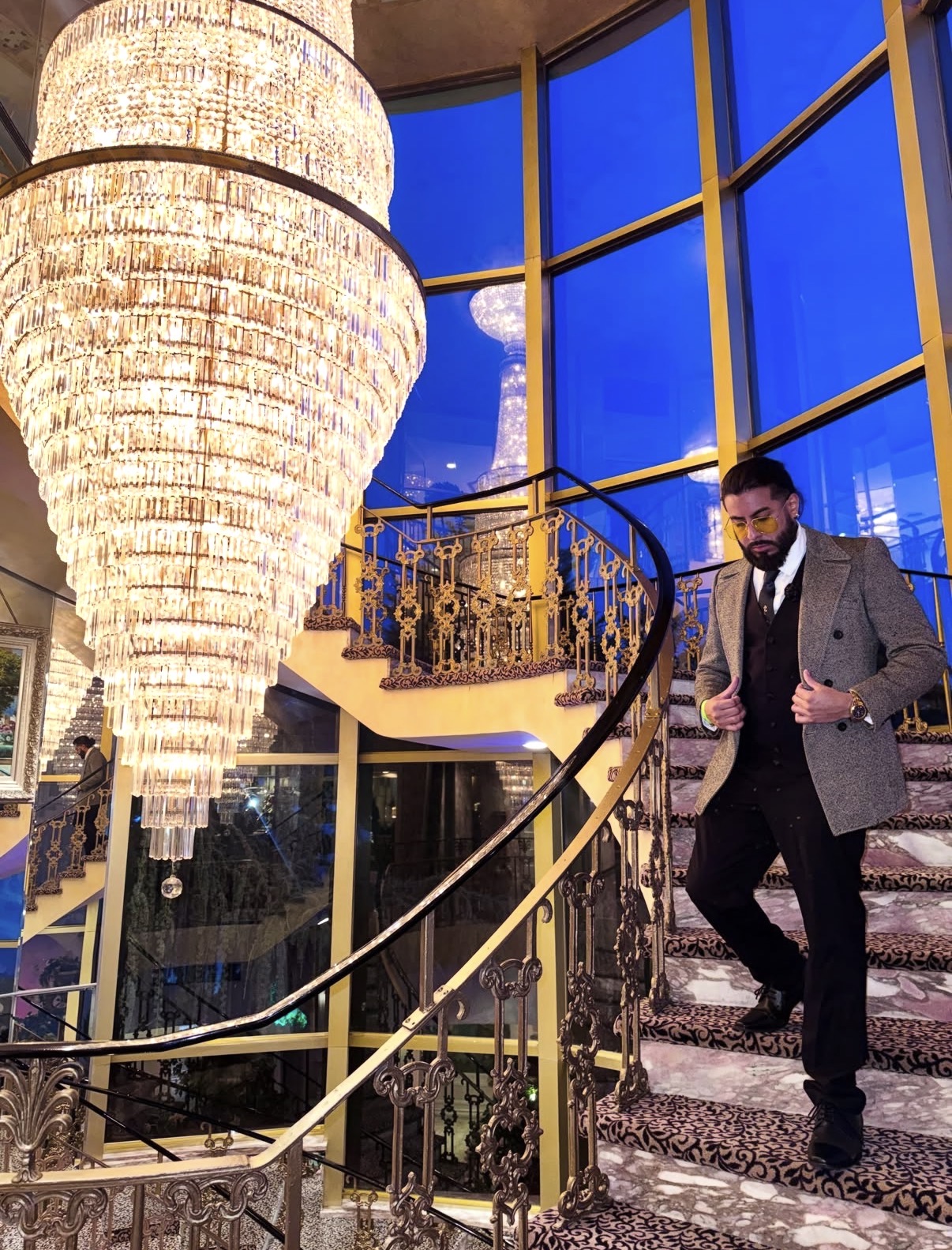 Richie in a tailored coat on a grand staircase under a crystal chandelier